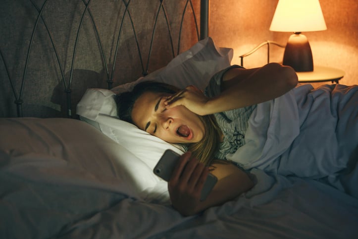 Une femme dans son lit baille devant son téléphone